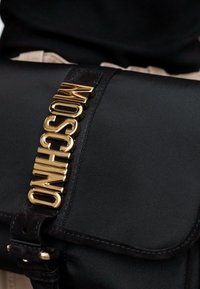 Must musta Moschino käekott kuldse logo tekstiga rihmal, hoitakse lähedal keha vastu musta ja beeži riiete taustal.
