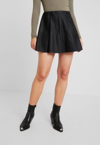 Jupe en cuir noir évasée avec une texture lisse et une taille haute. Portée avec un haut moulant vert olive et des bottines noires à talons pointus.