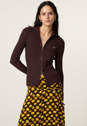 Vrouw met donker krullend haar draagt een bruine geribbelde trui met rits en een lange rok met een felgeel rozenbloemenpatroon.