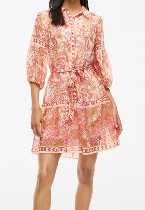 Vrouw draagt een jurk met een roze en oranje bloemenprint, 3/4 pofmouwen, knopen aan de voorkant en een gestrikte tailleband.