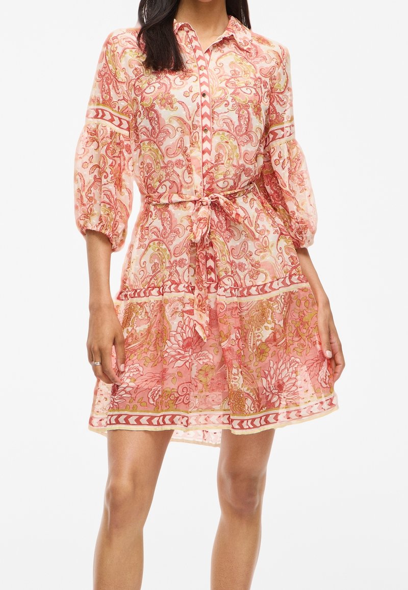Femme portant une robe à motifs floraux rose et orange avec des manches bouffantes 3/4, boutonnée devant, et une ceinture nouée à la taille.
