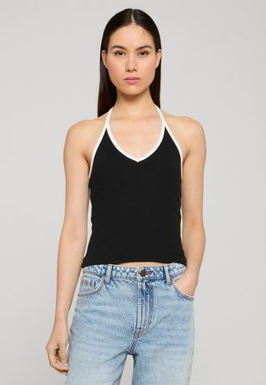 Femme aux longs cheveux noirs portant un haut dos-nu noir avec un bord blanc et un jean taille haute bleu clair, debout devant un fond uni.