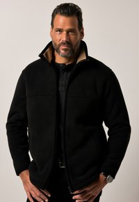 Chaqueta de forro polar negra con cierre de cremallera, cuello alto y forro beige. Presenta un pequeño logotipo frontal y una tapeta con botones. Tejido texturizado.