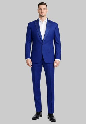 Homme debout face à l'avant, portant un costume bleu roi ajusté avec des revers brillants, une chemise blanche et des chaussures noires sur fond gris.