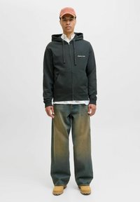 Sort hættejakke med lynlås og snore, hvidt tekst på brystet, parret med løse grønne denimjeans og brune støvler, båret over en hvid skjorte.