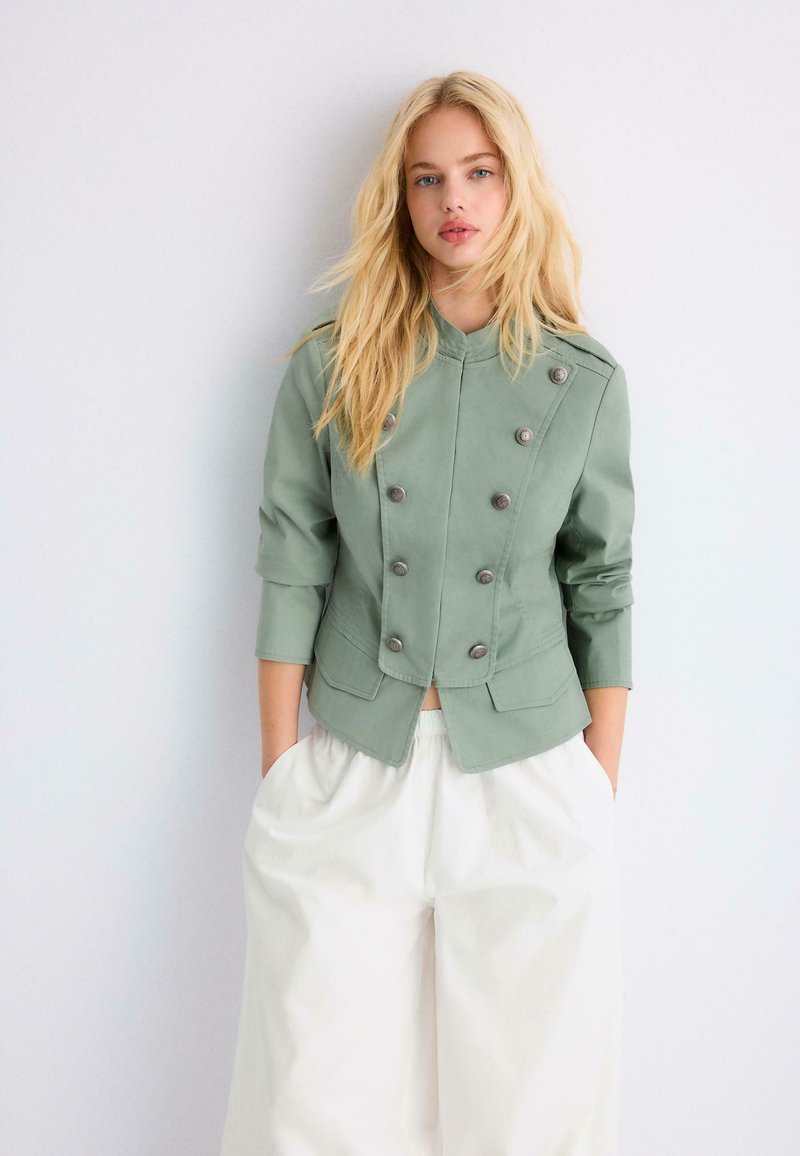 Jeune femme aux longs cheveux blonds portant une veste verte à double boutonnage et un pantalon large blanc, debout devant un mur clair uni.