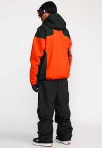 Veste de snowboard dans des couleurs orange vif et noire, dotée d'une capuche, de poignets ajustables, et accompagnée d'un pantalon noir ample et de gants.