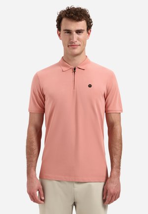 Man draagt een zalmroze polo met korte mouwen en een halve ritskraag en beige broek, staand tegen een witte achtergrond.