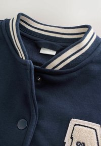 Marineblå varsityjakke med hvidstribet krave, knapper og en cremefarvet lap med teksturerede detaljer.