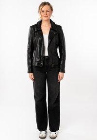 Giacca da motociclista in pelle nera con colletto a punta e chiusura zip, abbinata a jeans scuri e sneakers bianche. Texture liscia, design aderente.