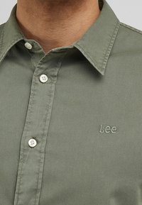 Close-up van een persoon die een olijfgroene button-up shirt draagt met een spitse kraag en witte knopen, geborduurd met het woord "Lee."