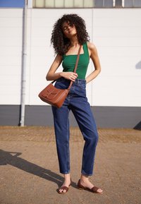 Débardeur vert côtelé, jean en denim bleu taille haute, sac bandoulière marron et sandales plates marron à texture tissée, debout sur une surface pavée.