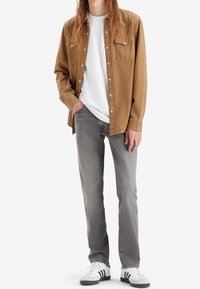 Camicia marrone con bottoni sopra una t-shirt bianca, jeans grigi e sneakers bianche con strisce nere. Caratteristiche includono due tasche sul petto e una vestibilità rilassata.