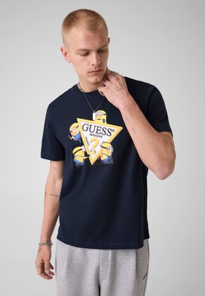 Jeune homme aux cheveux courts portant un t-shirt Guess Minions bleu marine et un pantalon de jogging gris clair, regardant vers le bas et touchant son cou.