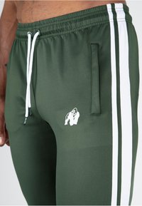 Groene sportieve joggers met witte zijstrepen, elastische tailleband, trekkoord en een klein wit gorilla-logo op de linker dij.