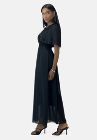 Robe maxi noire avec taille smockée, manches courtes volantées et tissu transparent. Associée à des sandales à talons hauts noires à lanières.