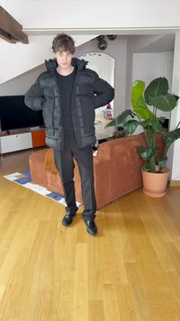 Juodas pūkinis švarkas su gobtuvu, su užtrauktuku priekyje ir banguotomis detalėmis, derinamas su juodomis kelnėmis ir batais, stovintis šviesioje vidaus erdvėje.