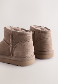 Paire de bottines beiges en daim avec doublure en shearling moelleuse et semelles en caoutchouc texturées, présentées de dos sur un fond neutre.