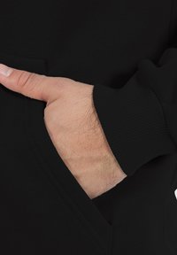 Sudadera negra con un bolsillo lateral, puños acanalados y una textura suave. Mano colocada en el bolsillo para resaltar el diseño.