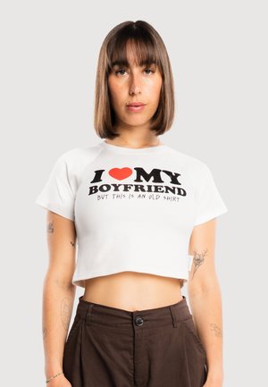 Młoda kobieta o średnio brązowych włosach nosi biały krótki t-shirt z napisem „I [heart] my boyfriend but this is an old shirt” oraz ciemne spodnie.