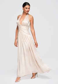 Femme aux cheveux foncés dans une robe de soirée fluide couleur ivoire à col halter et sandales à talons hauts, posant devant un fond clair uni.