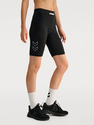 Model draagt zwarte knielange Hummel shorts met wit chevron-logo, witte sokken en zwarte sportschoenen tegen een effen achtergrond.