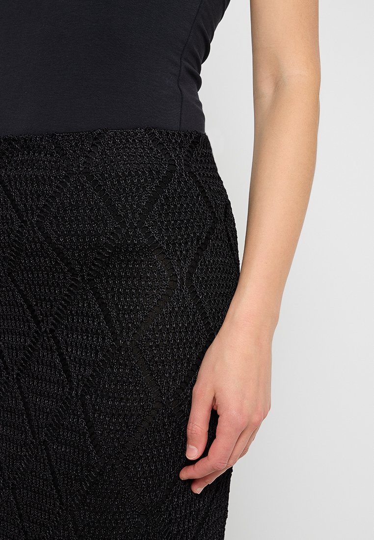 Schwarzer, strukturierter Rock mit einem rautenförmigen Muster, aus einem gewebten Stoff gefertigt. Eine Hand ruht sanft am Rand des Rocks.