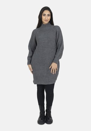 Kvinde står iført en grå ribstrikket rulneck-kjole, sorte leggings og sorte snørebåndsstøvler på en lys baggrund.