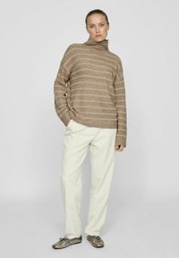 Bež svilen jopič s turtleneck ovratnikom in ohlapnim krojem, v kombinaciji z belimi hlačami in metalnimi natikači, postavljen na enotnem ozadju.