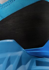 Tige de chaussure de sport bleu avec des accents texturés noirs, dotée d'un talon lisse et contourné. Les matériaux comprennent un tissu synthétique et du caoutchouc.