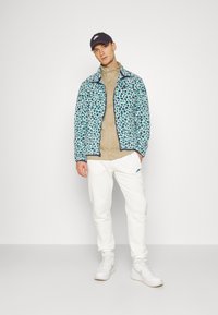 Jachetă cu imprimeu leopard bleu deschis, cu guler înalt, cămașă bej, pantaloni sport albi și adidași albi. O șapcă închisă completează ținuta.