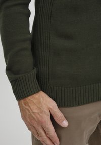Tmavo zelený pletený sveter s rebrovanými manžetami a lemom, zvýrazňujúci textúrovaný materiál a priliehavý strih. Ruka spočívajúca na svetlohnedej látke.