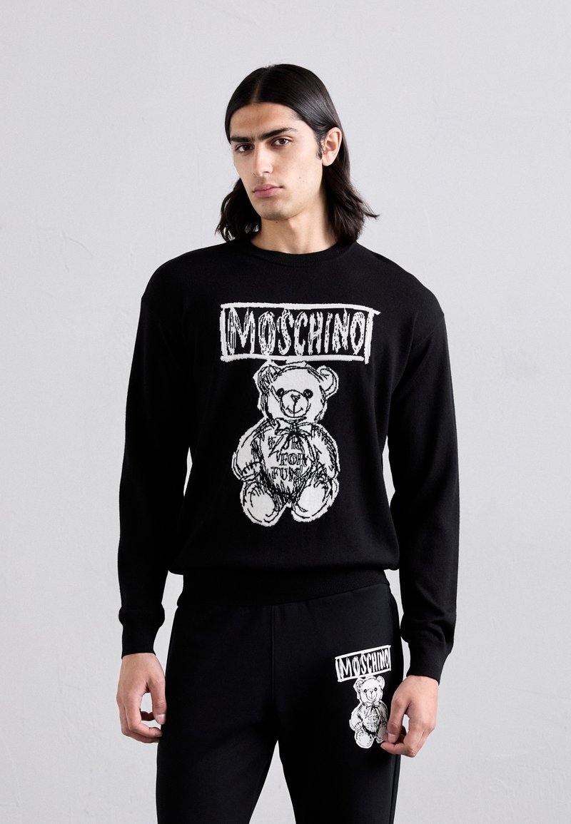El suéter negro presenta un gráfico de un oso con el texto "Moschino" encima y "Toy for Fun" debajo, combinado con pantalones negros a juego.