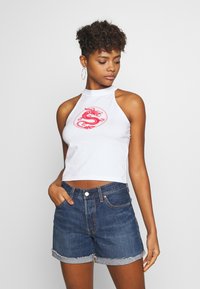 Hvit halterneck crop-top med rød dragegrafikk, kombinert med mørkeblå denimshorts med slitt kant. Modellen står med den ene hånden i lommen.
