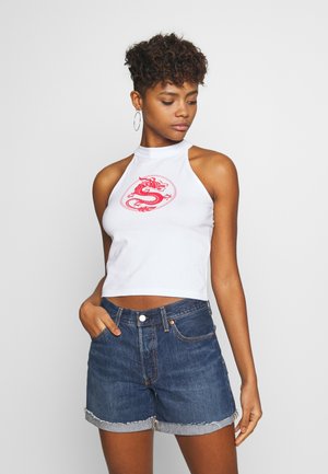 Hvit halterneck crop-top med rød dragegrafikk, kombinert med mørkeblå denimshorts med slitt kant. Modellen står med den ene hånden i lommen.