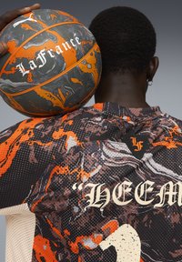 Basketball med sort, orange og grå marmorering, med teksten "La France". Spiller bærer trøje med abstrakt design og logo.
