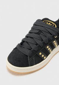 Melna zamšādas sporta apavi ar bieziem pelēkiem šņorēm, zelta akcentiem sānos un baltu gumijas zoli. Noapaļa purngala forma un teksturēta virsma.