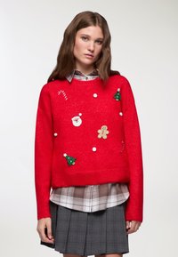 Czerwony sweter robiony na druty z świątecznymi ozdobami: Mikołaj, piernikowy ludzik, choinki i pompony. Nałożony na koszulę w kratę i spódnicę.