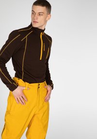 Hnedá mikina na zips so žltými akcentami, skombinovaná s jasne žltými nohavicami so sponou a vreckami. Hladké, funkčné materiály.