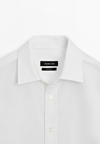 Camicia bianca con colletto classico, chiusura frontale con bottoni a perla e un'etichetta del marchio discreta sul collo.