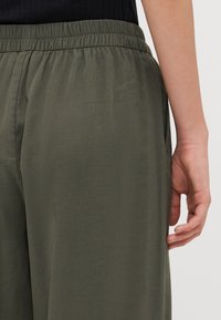 Olivengrønne, lette bukser med elastisk talje; har sidelommer og en glat tekstur; beskåret længde vist fra siden.