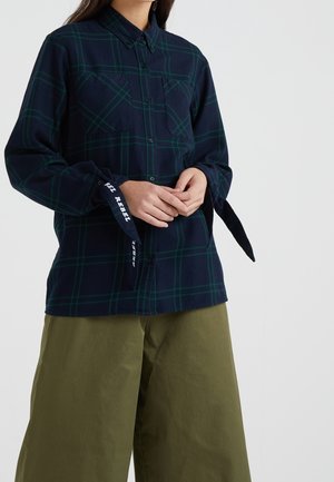 Kobieta w granatowo-zielonej koszuli w kratę z napisem „BE REBEL” na mankietach i oliwkowo-zielonych szerokich spodniach, stojąca z rękami złączonymi.