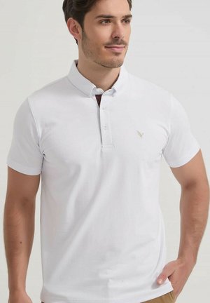 Homme aux cheveux courts et foncés portant un polo blanc à manches courtes avec un col boutonné et un petit logo sur la poitrine, debout devant un fond uni.