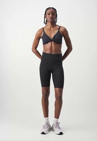 Fekete sportcrop top csavart elülső dizájnnal, hozzáillő fekete kerékpáros rövidnadrággal. Felnőtt modell semleges háttér előtt áll, sportcipőt viselve.