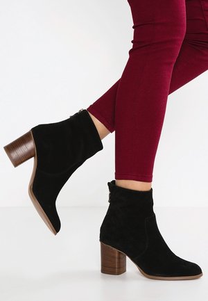 Bottines en daim noir avec un bout arrondi et un talon bloc en bois de 7 cm. Elles possèdent une fermeture éclair sur le côté extérieur et une texture douce.