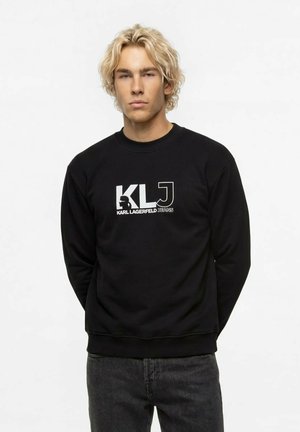 Mladý muž s blond vlasmi, ktorý nosí čiernu mikinu s logom "KLJ Karl Lagerfeld Jeans" a tmavé rifle, stojaci na bielom pozadí.