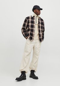 Geruite shirt in donkerblauw, bruin en beige over een crèmekleurige zip-necksweater, gecombineerd met losse beige broek en zwarte gevechtslaarzen.