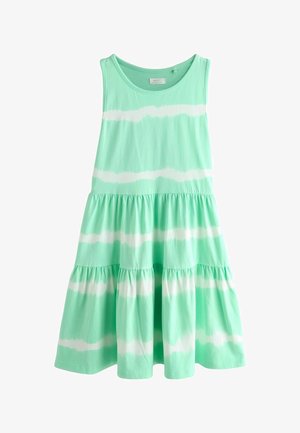 Hosszú, göndör hajú fiatal lány, aki ujjatlan, mentazöld és fehér csíkos ruhát visel, valamint fehér pántos szandált, egyik lábát előre nyújtva áll.