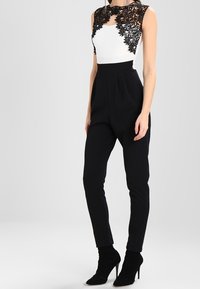 Bodysuit en dentelle noire avec panneaux en dentelle, haut blanc, pantalons noirs taille haute et bottines noires, présentant une silhouette ajustée et une texture lisse.