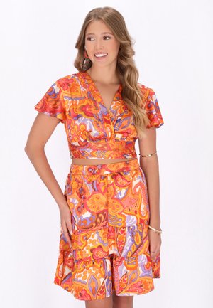 Tânără cu păr lung și ondulat, purtând un top scurt colorat cu model floral în nuanțe de portocaliu, roz și mov, asortat cu o fustă în același stil.